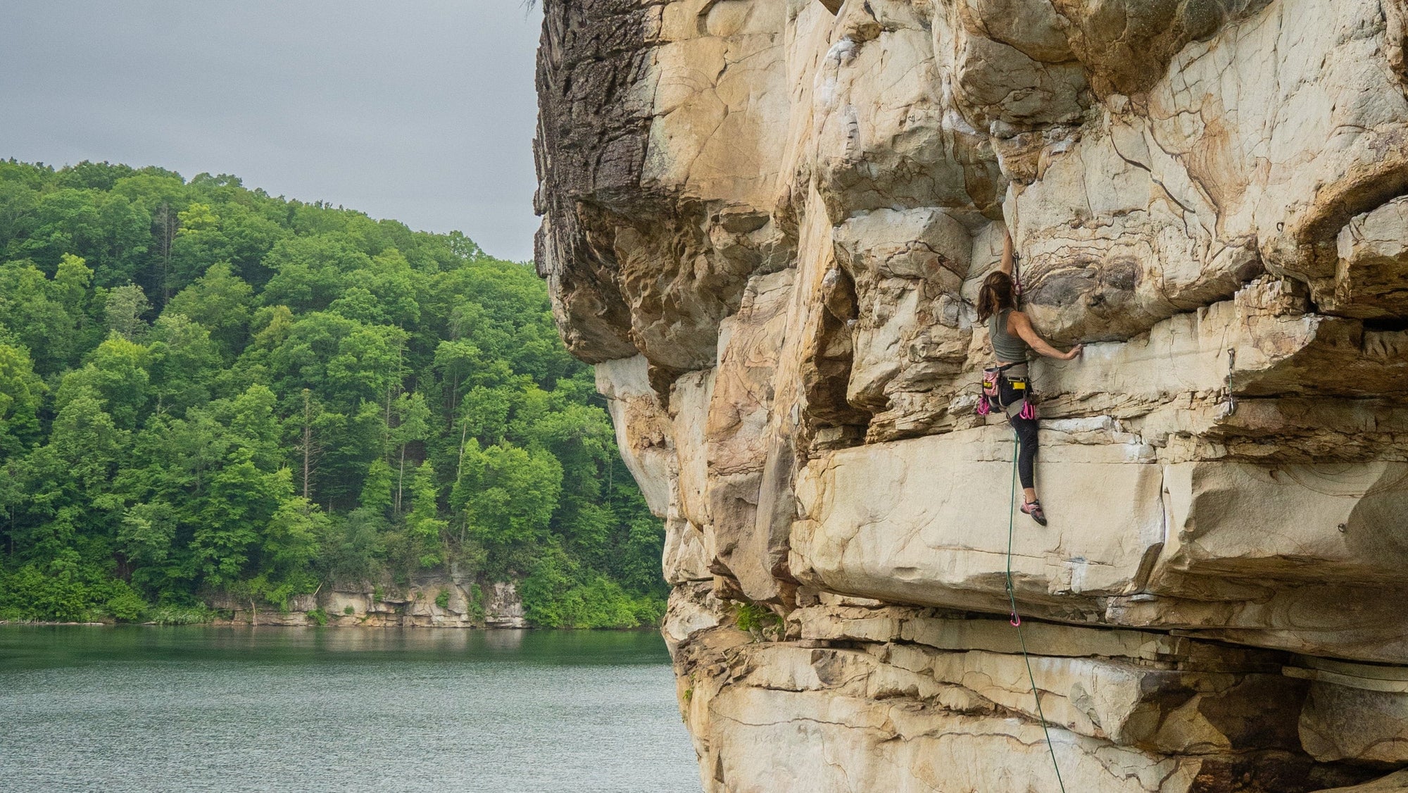 Person climbing a rocky cliff with a lake and trees in the background